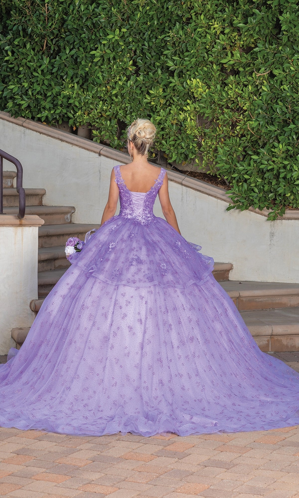 Wearing the Quinceanera Dress 1702 By Dancing Queen, a person stands outdoors on a stone pathway in a floor-length lavender ball gown with an elegant lace cape, facing away amidst greenery and stairs.